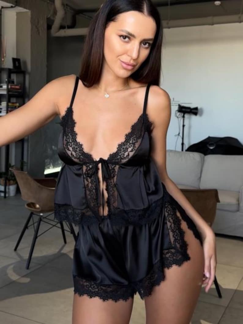 May include: A black two-piece pajama set consisting of a camisole top and matching shorts. The camisole features thin shoulder straps, a deep V-neckline, and black lace trim. The shorts have a high-cut leg design with matching black lace trim.