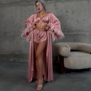 May include: A pink satin robe with feather trim on the sleeves. The robe is worn over a matching pink satin bra and shorts set.