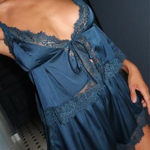 May include: A dark teal satin pajama set consisting of a camisole and shorts. The camisole features thin shoulder straps, a deep V-neckline with intricate lace trim, and a center tie detail. The matching shorts have a wide lace hem.