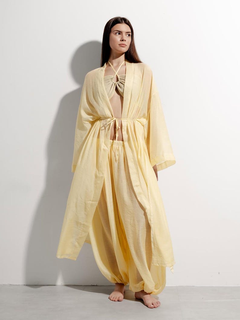 May include: A pale yellow three-piece ensemble: a halter top, harem trousers, and a long, open-front robe. The robe features wide sleeves and a tie at the waist. The trousers have gathered ankles. The outfit is crafted from a lightweight, flowing material.