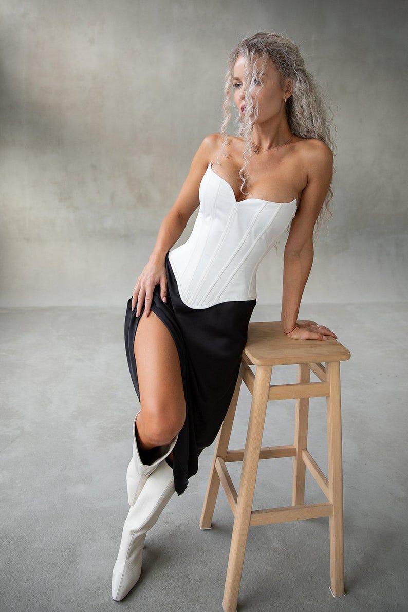 May include: A white corset with a sweetheart neckline is paired with a black skirt featuring a high slit. The model is wearing white boots and leaning on a wooden stool. The background is a neutral gray.