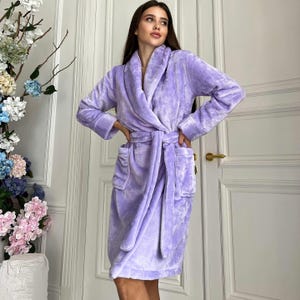 Peut inclure: Un peignoir de couleur lavande avec un col châle et une ceinture assortie. La robe a des manches longues et des poches. Le tissu en peluche semble confortable et chaud. La robe arrive au genou et convient pour se prélasser.
