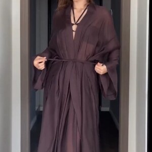 May include: A long, flowing, dark brown robe with a tie waist and wide sleeves. The robe has a deep V-neckline and is made of a sheer, lightweight fabric. The model is standing in a hallway.