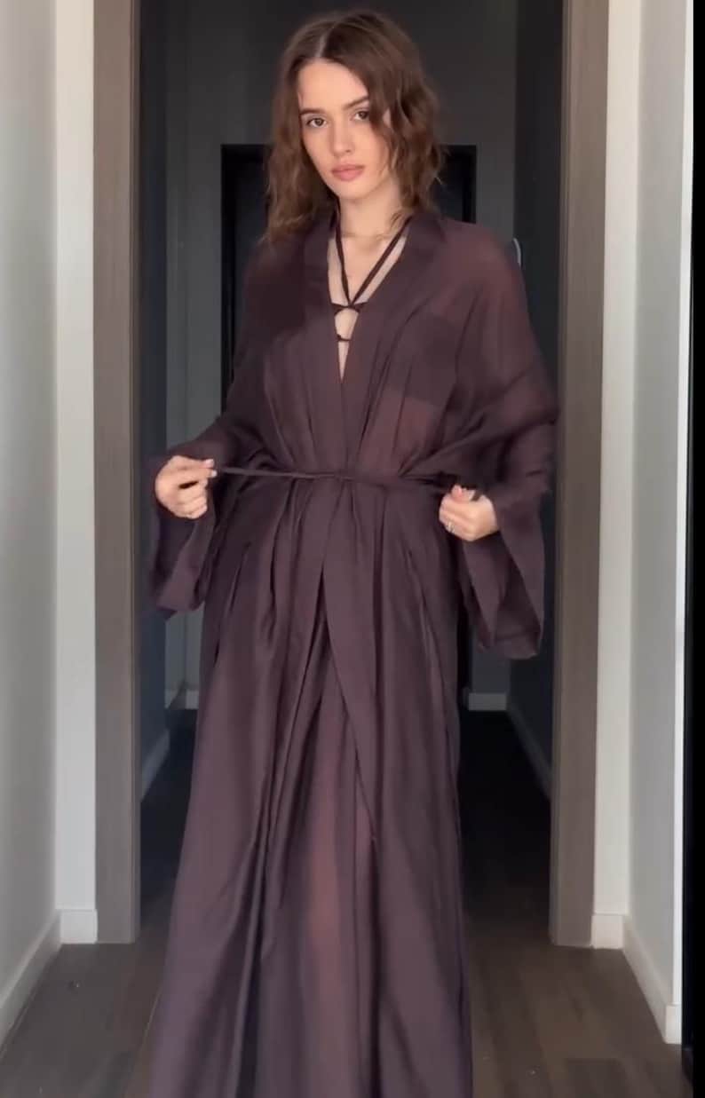 May include: A long, flowing, dark brown robe with a tie waist and wide sleeves. The robe has a deep V-neckline and is made of a sheer, lightweight fabric. The model is standing in a hallway.