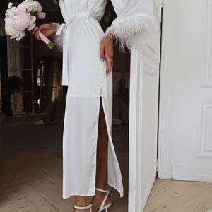 May include: A white satin robe with feather trim on the sleeves and a matching belt. The robe has a high slit on the side and is paired with white heeled sandals. A bouquet of pink and white flowers is held in the hand.