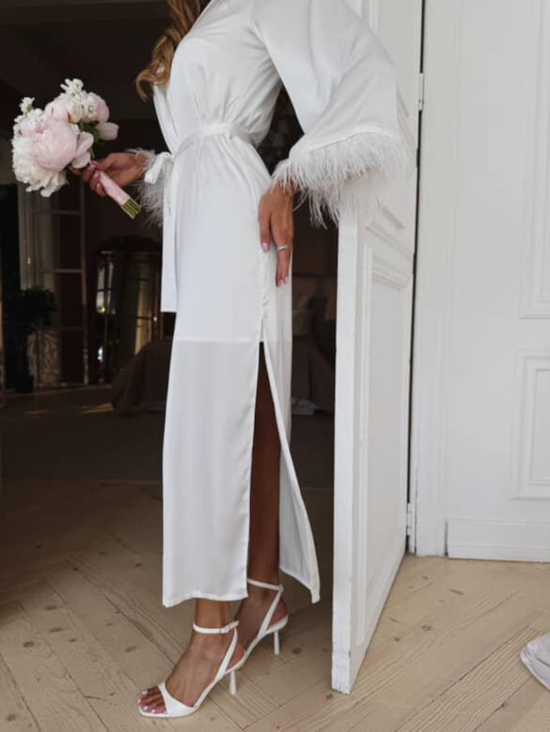 May include: A white satin robe with feather trim on the sleeves and a matching belt. The robe has a high slit on the side and is paired with white heeled sandals. A bouquet of pink and white flowers is held in the hand.