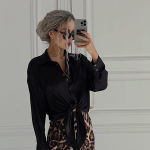 May include: A woman wearing a black satin shirt tied at the waist over a brown and black leopard print skirt. She is wearing sunglasses and has her hair pulled back in a bun.