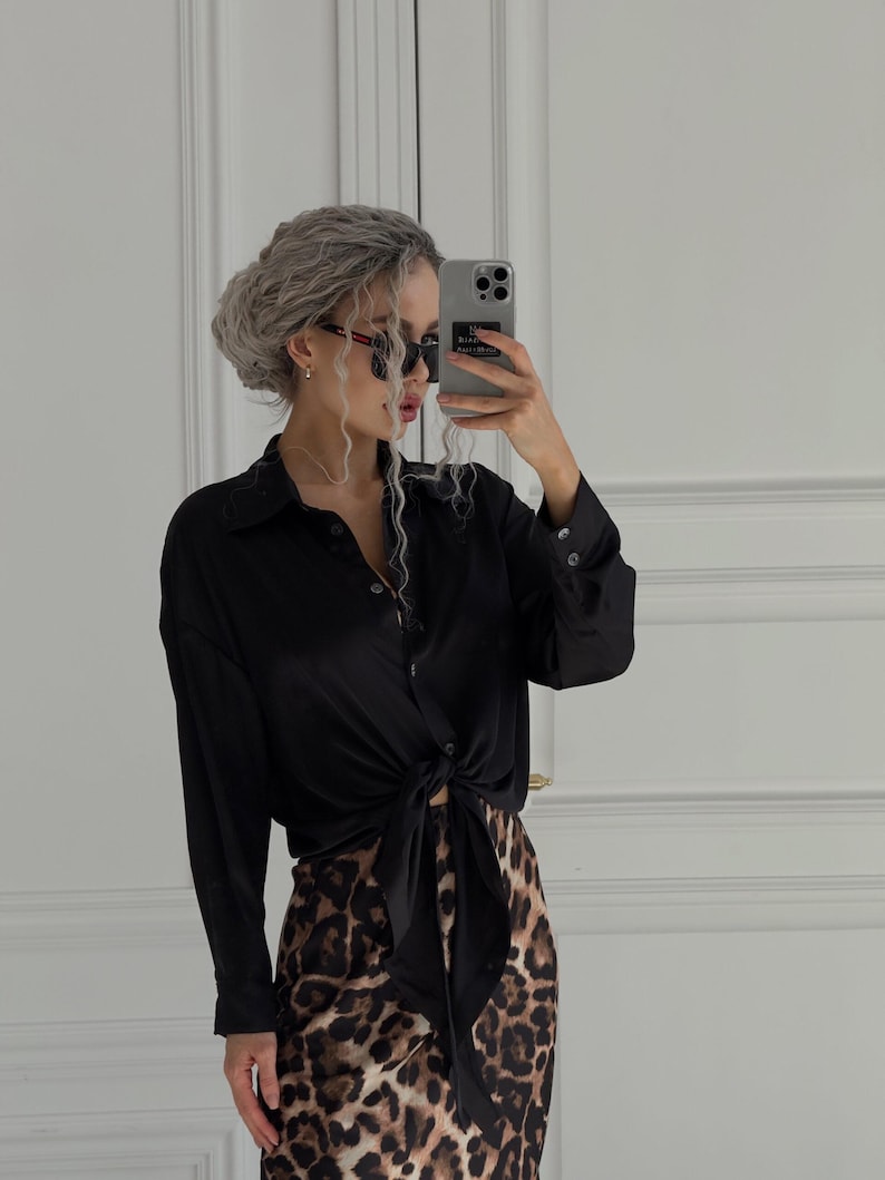 May include: A woman wearing a black satin shirt tied at the waist over a brown and black leopard print skirt. She is wearing sunglasses and has her hair pulled back in a bun.