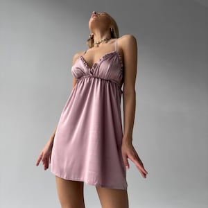 May include: A light mauve satin slip dress with spaghetti straps and ruffled trim. The dress has a gathered bodice and a flared skirt. The model is wearing a gold necklace. The dress is a mini length.