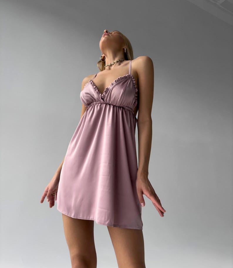 May include: A light mauve satin slip dress with spaghetti straps and ruffled trim. The dress has a gathered bodice and a flared skirt. The model is wearing a gold necklace. The dress is a mini length.