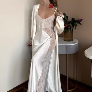 May include: A white satin slip dress with lace detailing and a matching long robe. The dress features a V-neck and a flowing silhouette. A red flower is held in the hand.