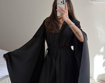 May include: A solid black floor-length robe featuring wide, dramatic bell sleeves and a matching fabric tie belt at the waist. The garment is crafted from a smooth, slightly lustrous material that drapes loosely, creating a clean and structured silhouette.