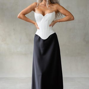 May include: A white, strapless corset top paired with a long, black skirt. The corset has a sweetheart neckline and structured boning. The skirt flows to the floor. The model is wearing white boots. The outfit is set against a neutral background.