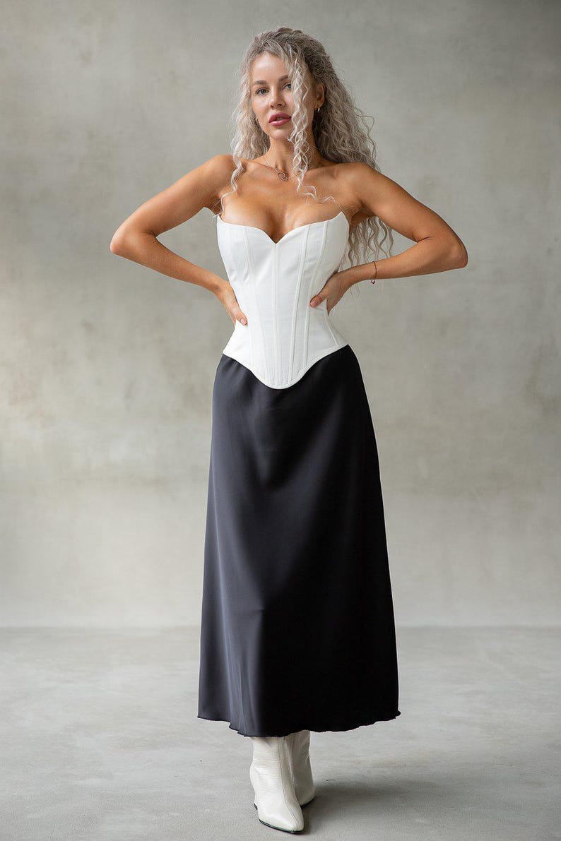 May include: A white, strapless corset top paired with a long, black skirt. The corset has a sweetheart neckline and structured boning. The skirt flows to the floor. The model is wearing white boots. The outfit is set against a neutral background.