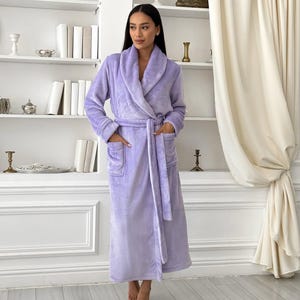 May include: A plush, lavender-colored bathrobe featuring a wide shawl collar, long sleeves, and two front patch pockets. The robe is secured at the waist with a matching fabric tie belt, creating a wrap-style closure that extends to mid-calf length.