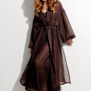 May include: A long, flowing brown robe with wide sleeves and a tie waist. The robe is made of a sheer fabric and is worn over a dark top. The model is standing in front of a white wall.