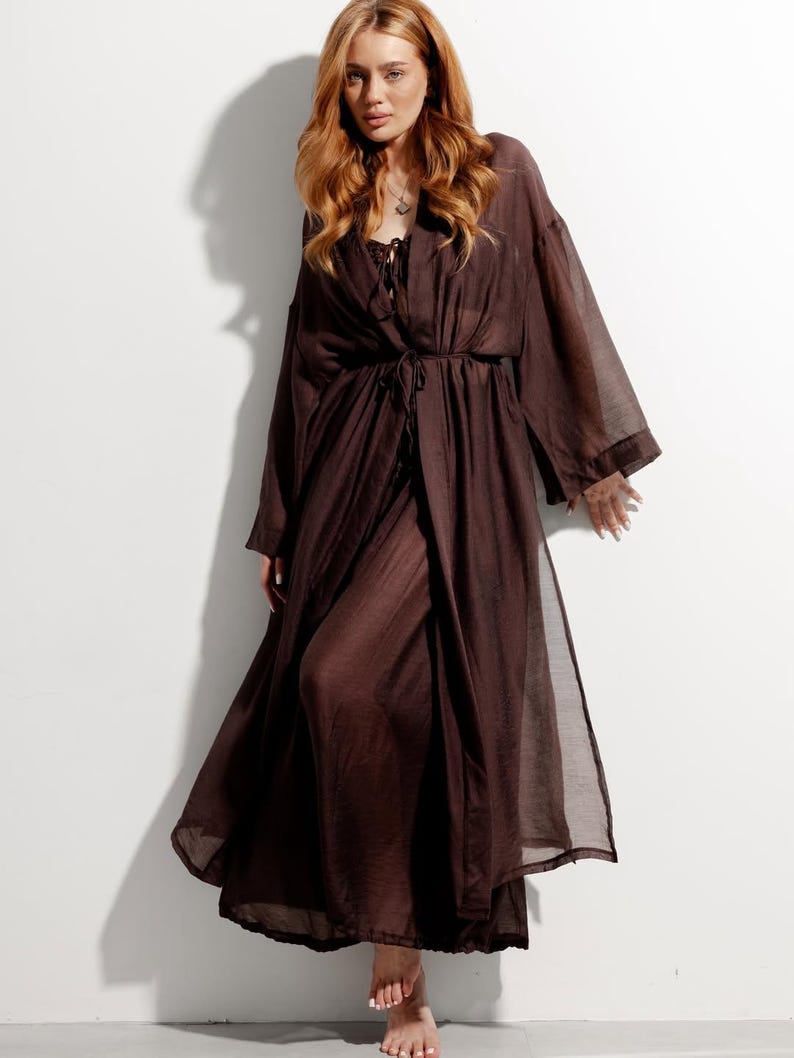 May include: A long, flowing brown robe with wide sleeves and a tie waist. The robe is made of a sheer fabric and is worn over a dark top. The model is standing in front of a white wall.