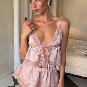 May include: A pale pink satin pajama set consisting of a camisole and shorts. The camisole features a deep V-neckline with delicate lace trim, thin shoulder straps, and a center front tie closure. The matching shorts have a scalloped lace hem.