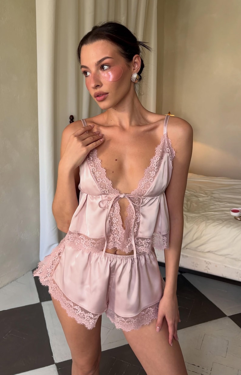 May include: A pale pink satin pajama set consisting of a camisole and shorts. The camisole features a deep V-neckline with delicate lace trim, thin shoulder straps, and a center front tie closure. The matching shorts have a scalloped lace hem.