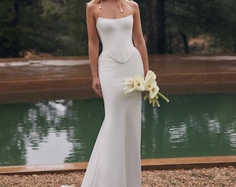May include: A floor-length white strapless gown featuring a structured corset-style bodice with vertical paneling and a smooth, fitted skirt that flares slightly at the hem. The garment is paired with a multi-strand pearl choker necklace.