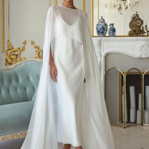 May include: A floor-length white dress featuring a sleeveless slip-style bodice with a scoop neckline and a sheer, flowing cape overlay that drapes from the shoulders to the floor, creating a layered silhouette with a clean, solid white finish.