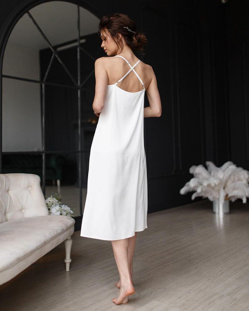 May include: A white slip dress with thin straps that cross in the back. The dress is made of a silky fabric and has a simple, elegant design.