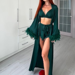 Puede incluir: Una bata de satén verde con ribetes de plumas en las mangas. La bata está abierta y se pueden ver los pantalones cortos y el sujetador a juego debajo. La persona lleva zapatillas de pelo rojo.