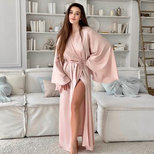 May include: A long, pink satin robe with a tie belt and wide sleeves. The robe is open at the front, revealing a glimpse of the model's legs.