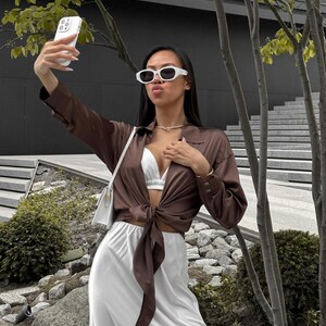 May include: A woman wearing a brown satin shirt tied at the waist over a white satin skirt. She is wearing white sunglasses and a white bra. She is holding a white iPhone with a white case and taking a selfie.