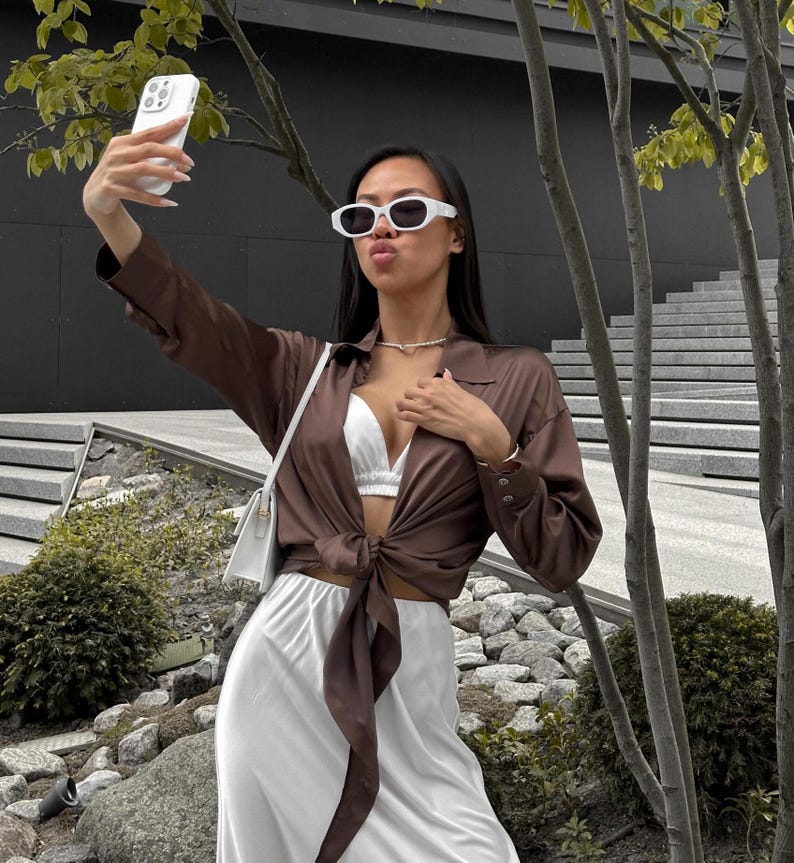 May include: A woman wearing a brown satin shirt tied at the waist over a white satin skirt. She is wearing white sunglasses and a white bra. She is holding a white iPhone with a white case and taking a selfie.