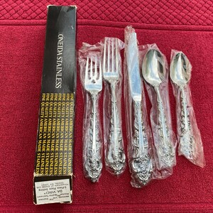 Könnte beinhalten: Ein Set Da Vinci Oneida Edelstahl-Besteck, einschließlich Gabeln, Messern und Löffeln, noch in der Originalverpackung. Die Schachtel ist schwarz mit goldfarbenen Buchstaben. Das Besteck hat ein detailliertes Blumendesign.