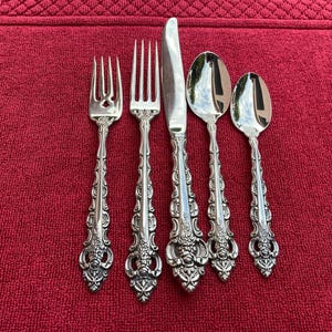 Könnte beinhalten: Ein Set aus fünf verzierten silberfarbenen Besteckteilen, darunter Gabeln, ein Messer und Löffel, angeordnet auf einer roten strukturierten Oberfläche. Das Besteck weist detaillierte Blumen- und Scrollmuster auf den Griffen auf.