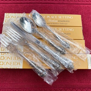 Può includere: Un set di posate nuove in acciaio inossidabile, tra cui forchette, cucchiai e coltelli, è confezionato in plastica trasparente. Le posate sono esposte sopra diverse scatole con la scritta "ONEIDA" e "PLACE SETTING".
