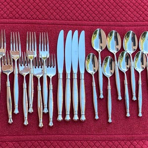 Op de afbeelding: Een set zilverkleurig bestek, inclusief vorken, messen en lepels, gerangschikt op een rode, getextureerde ondergrond. Het bestek heeft een eenvoudig, elegant ontwerp met een gepolijste afwerking, geschikt voor formele diners.