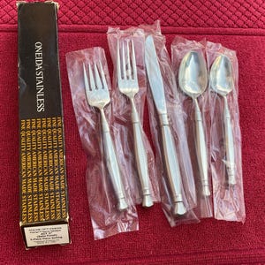 May include: A new 5-piece stainless steel flatware set, still in its original packaging, includes forks, knives, and spoons. The box reads "ONEIDA STAINLESS" and "FINE QUALITY AMERICAN MADE STAINLESS". The set is displayed on a red textured surface.