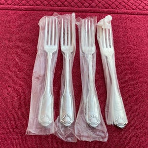 Op de afbeelding: Vier zilveren vorken, elk afzonderlijk verpakt in doorzichtig plastic. De vorken hebben een decoratief schelpontwerp op het handvat. De vorken zijn gerangschikt op een rode, getextureerde ondergrond. Geschikt voor formele diners.
