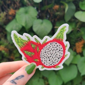 May include: A sticker featuring a detailed embroidered design of a dragon fruit. The sticker showcases the fruit's vibrant red skin, green leafy accents, and the white flesh dotted with black seeds. The sticker is held against a backdrop of green foliage.