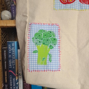 May include: A white canvas tote bag with a blue and white gingham patch featuring a green broccoli illustration. The bag has red and yellow stitching around the patch. The bag is sitting on a shelf with books.