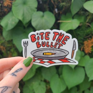 May include: A sticker with the text "Bite the Bullet" in red and black. The sticker features a cartoon image of a plate with a bullet on it, a fork, and a knife. The background is green.
