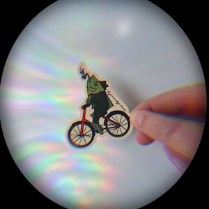 May include: A sticker featuring a fish riding a red bicycle. The fish has green scales and is wearing a hat. The sticker has the text "@janandjo.studio". The background is white with rainbow light reflections.