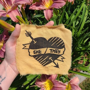 May include: A tan fabric patch with a black heart pierced by an arrow. The heart has diagonal lines and a banner reading "SHE / THEY". The patch has a frayed edge. Flowers with pink petals and yellow centers are in the background.
