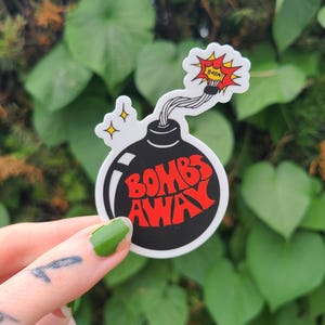 May include: A black and white sticker in the shape of a bomb with the words "BOMBS AWAY" in red. The sticker has a white outline and a red and yellow explosion graphic. Two yellow stars are on the left side of the sticker.
