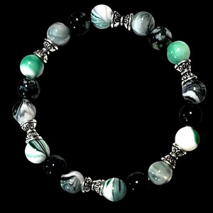 May include: A beaded bracelet featuring a variety of round beads in shades of green, white, and black. The beads are separated by ornate silver-colored metal spacers. The bracelet is displayed against a black background.