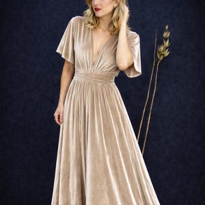 Könnte beinhalten: Ein bodenlanges, beigefarbenes Samtkleid mit tiefem V-Ausschnitt und kurzen, ausgestellten Ärmeln. Das Kleid hat eine taillierte Taille und einen fließenden Rock. Das Model steht vor einem dunklen Hintergrund.
