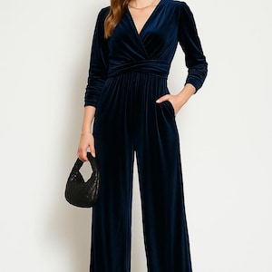 May include: A navy blue velvet jumpsuit with a wrap-style bodice and wide-leg trousers. The long-sleeved jumpsuit features a V-neckline and pockets. A small black quilted handbag is held in the left hand. The model is wearing gold sandals.