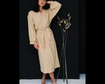 May include: A long, beige robe with a tie belt and pockets. The robe is made of a soft, lightweight fabric and has a relaxed fit. The model is wearing the robe with a simple necklace and pink shoes.