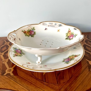 May include: A white porcelain bowl with a gold rim and floral design. The bowl has three legs and a strainer bottom. It is sitting on a matching plate with a floral design.