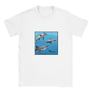 以下が含まれることがあります： 白いTシャツ。四角いグラフィックで、青い水中で泳ぐサメが描かれています。ハンマーヘッドシャーク、ホホジロザメ、ダイバーが含まれています。アーティストのサインが下部にあります。