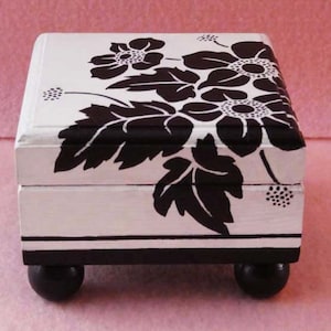 May include: A white wooden box with black floral designs and black feet. The box has a hinged lid and is decorated with a black and white floral pattern.