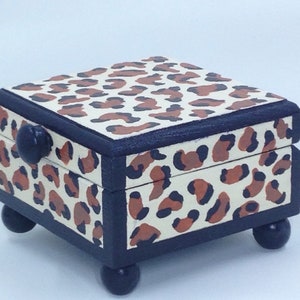 May include: A small wooden box with a leopard print design. The box has a black trim and a black knob. The box is sitting on four black feet.
